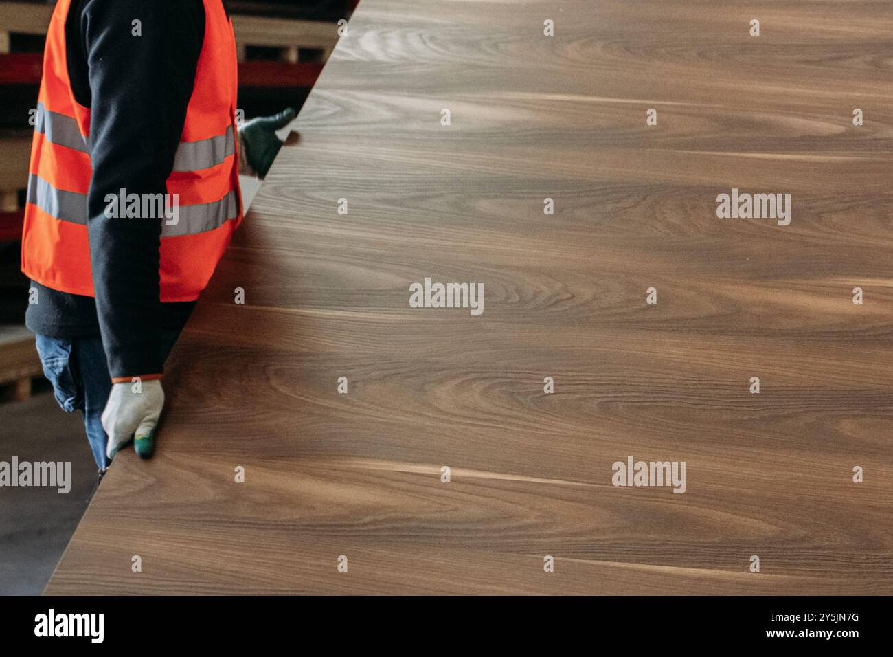 Un lavoratore che indossa un giubbotto di sicurezza maneggia con cura un grande pezzo di legno in un impianto di produzione di mobili. Foto Stock