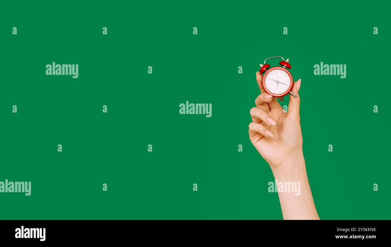 Sbrigati. Promemoria orario. Offerta Last Minute. Mano femminile che mostra l'ora su una sveglia rossa miniaturizzata d'epoca isolata su sfondo verde a spazio vuoto. Foto Stock
