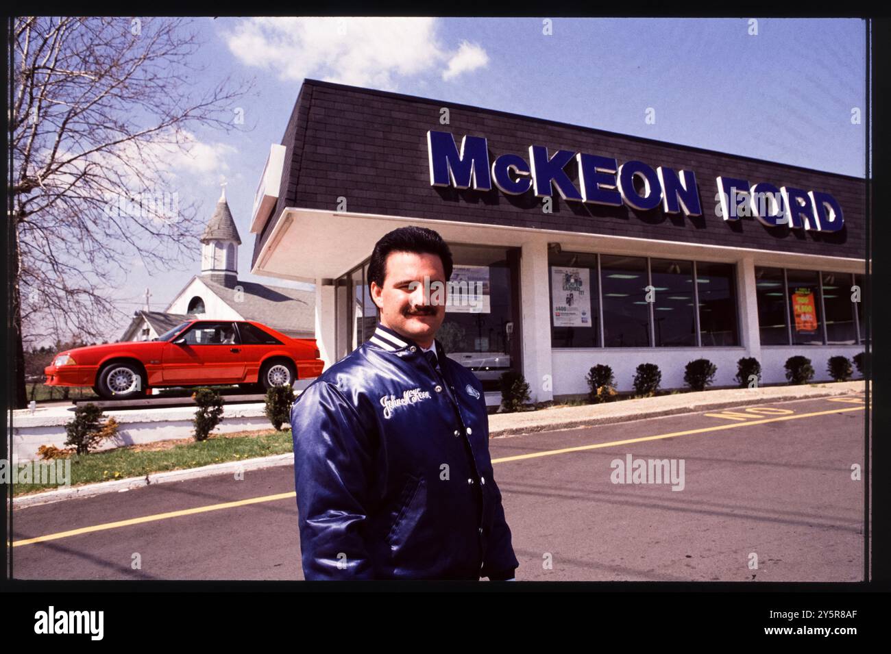 Un orgoglioso venditore si trova fuori McKeon Ford a Trenton, New Jersey, alla fine degli anni '1980, indossando una giacca da concessionaria. Dietro di lui, una Ford Mustang rossa, uno dei modelli iconici di Ford, viene esposta in modo prominente, evidenziando l'attenzione della concessionaria sui veicoli ad alte prestazioni. Foto Stock