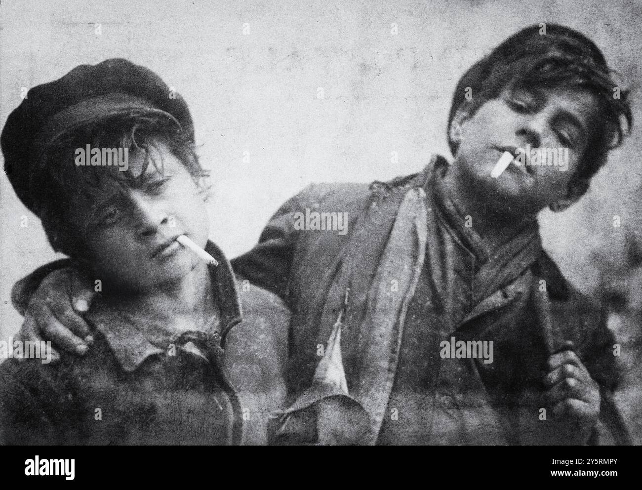 Due ragazzi stanno fianco a fianco, ognuno fumando una sigaretta, riflettendo uno spirito di cameratismo e di giovani spensierati nei primi anni '1900 Il loro abbigliamento casual richiama un background di classe operaia. Foto Stock