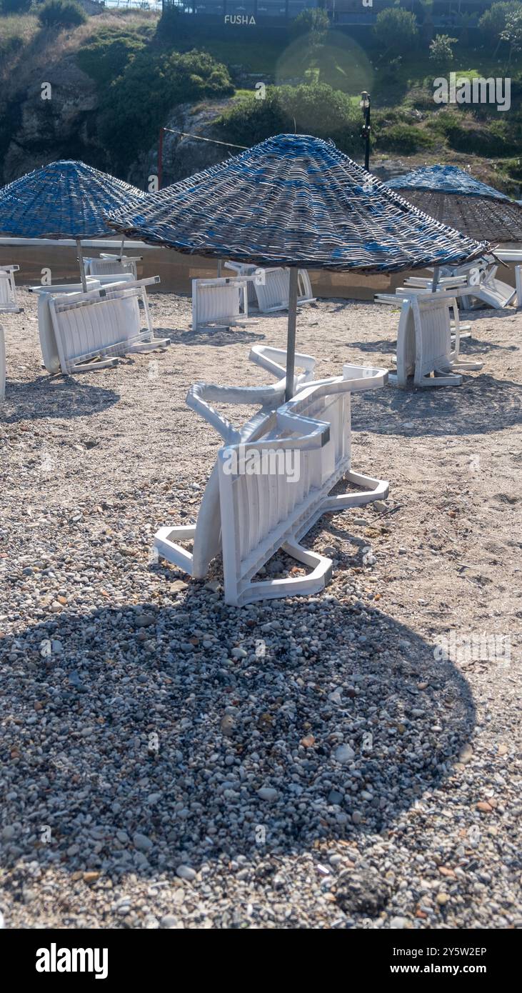 La luce del sole proietta ombre su sdraio e ombrelloni impilati in una tranquilla posizione sul mare, circondata da scogliere rocciose Foto Stock