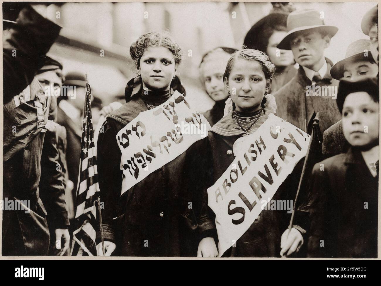 Protesta contro il lavoro minorile in una parata di lavoro. STATI UNITI. 1° maggio 1909 la fotografia mostra un ritratto a mezza lunghezza di due ragazze che indossano striscioni con lo slogan "ABOLIRE LA SCHIAVITÙ INFANTILE!!" In inglese e yiddish, una porta bandiera americana; gli spettatori si trovano nelle vicinanze. Probabilmente durante la parata del 1° maggio 1909 a New York. Foto Stock