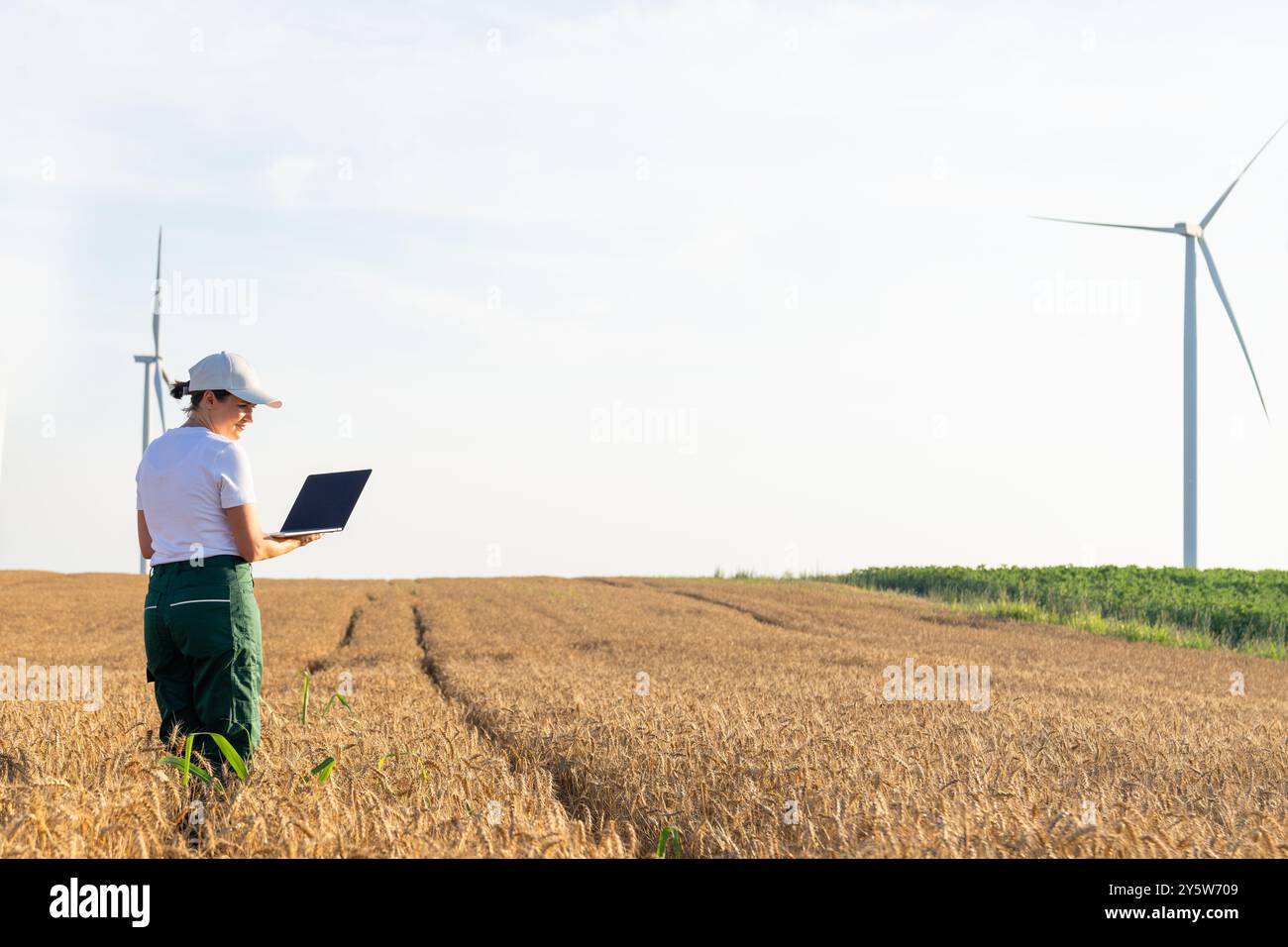 Donna agricoltrice che indossa un berretto bianco e una t-shirt con laptop sul campo agricolo al tramonto. Turbine eoliche sullo sfondo... Foto Stock