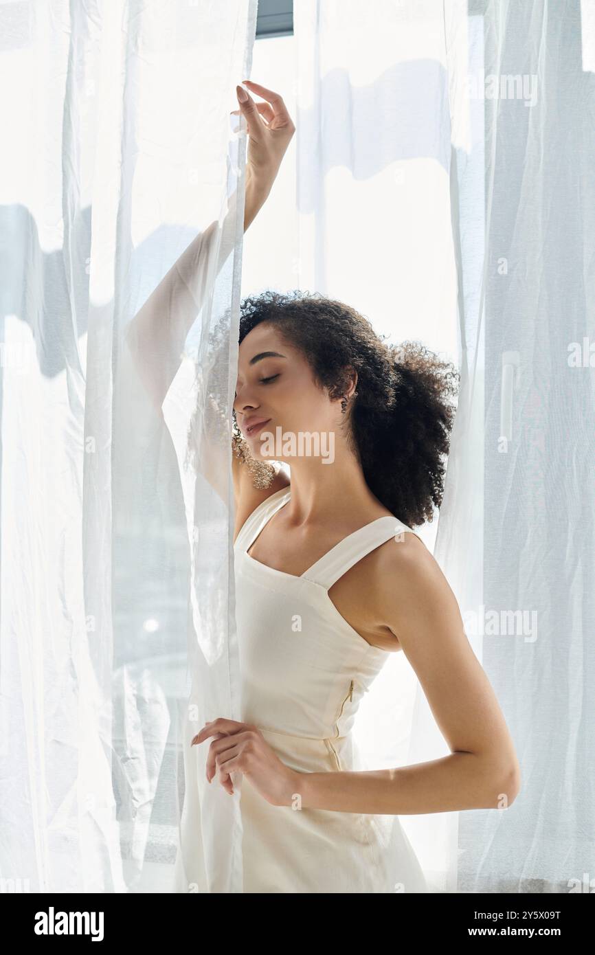 Una donna alla moda sta in piedi con tende che scorrono, immergendosi nella calda luce del sole mentre posa elegantemente. Foto Stock