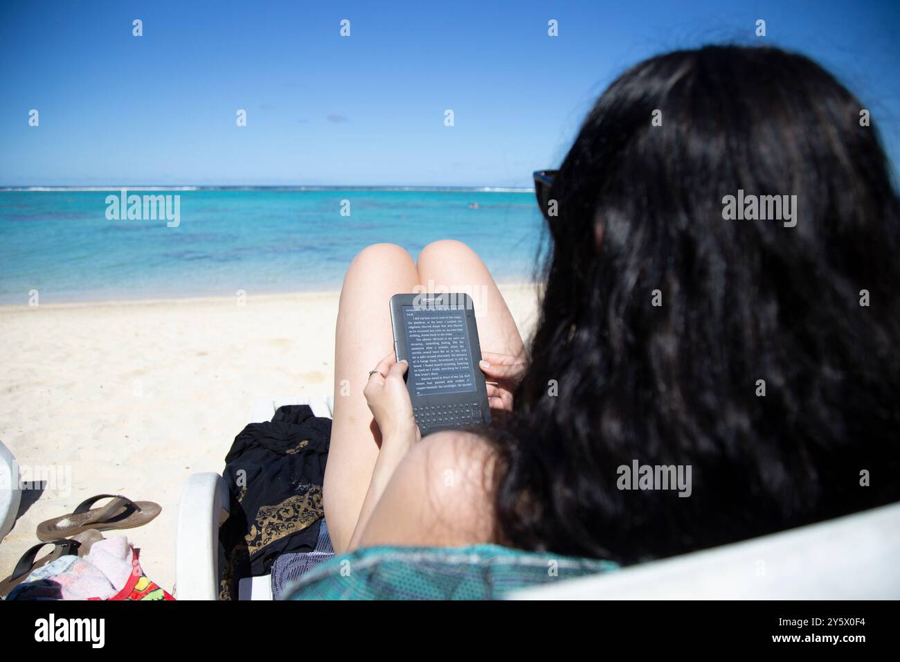 Donna che legge un eBook su una spiaggia sabbiosa con un oceano azzurro cristallino sullo sfondo, Titikaveka, Rarotonga, Isole Cook Foto Stock