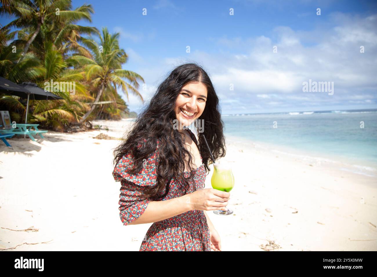 Donna sorridente che regge un bicchiere di bevanda tiepida su una spiaggia soleggiata con palme e cielo blu sullo sfondo, Titikaveka, Rarotonga, Isole Cook Foto Stock