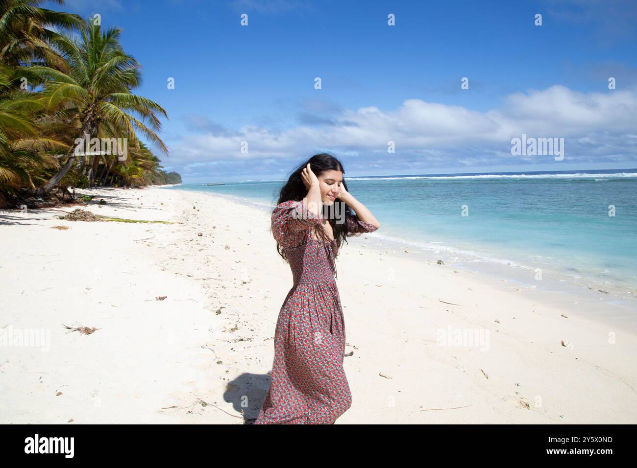 Una donna con un abito floreale sorride mentre cammina lungo una spiaggia tropicale con palme e oceano sullo sfondo, Titikaveka, Rarotonga, Isole Cook Foto Stock