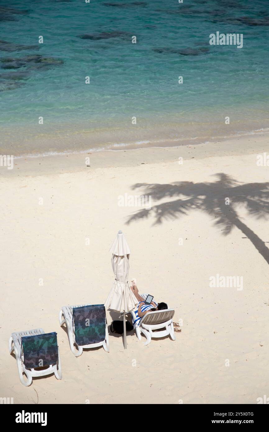 Donna che legge un e-book sulla spiaggia, lettini bianchi e ombrellone chiuso, una palma getta un'ombra vicino alle acque cristalline turchesi, Titikaveka, Rarotonga, Isole Cook Foto Stock