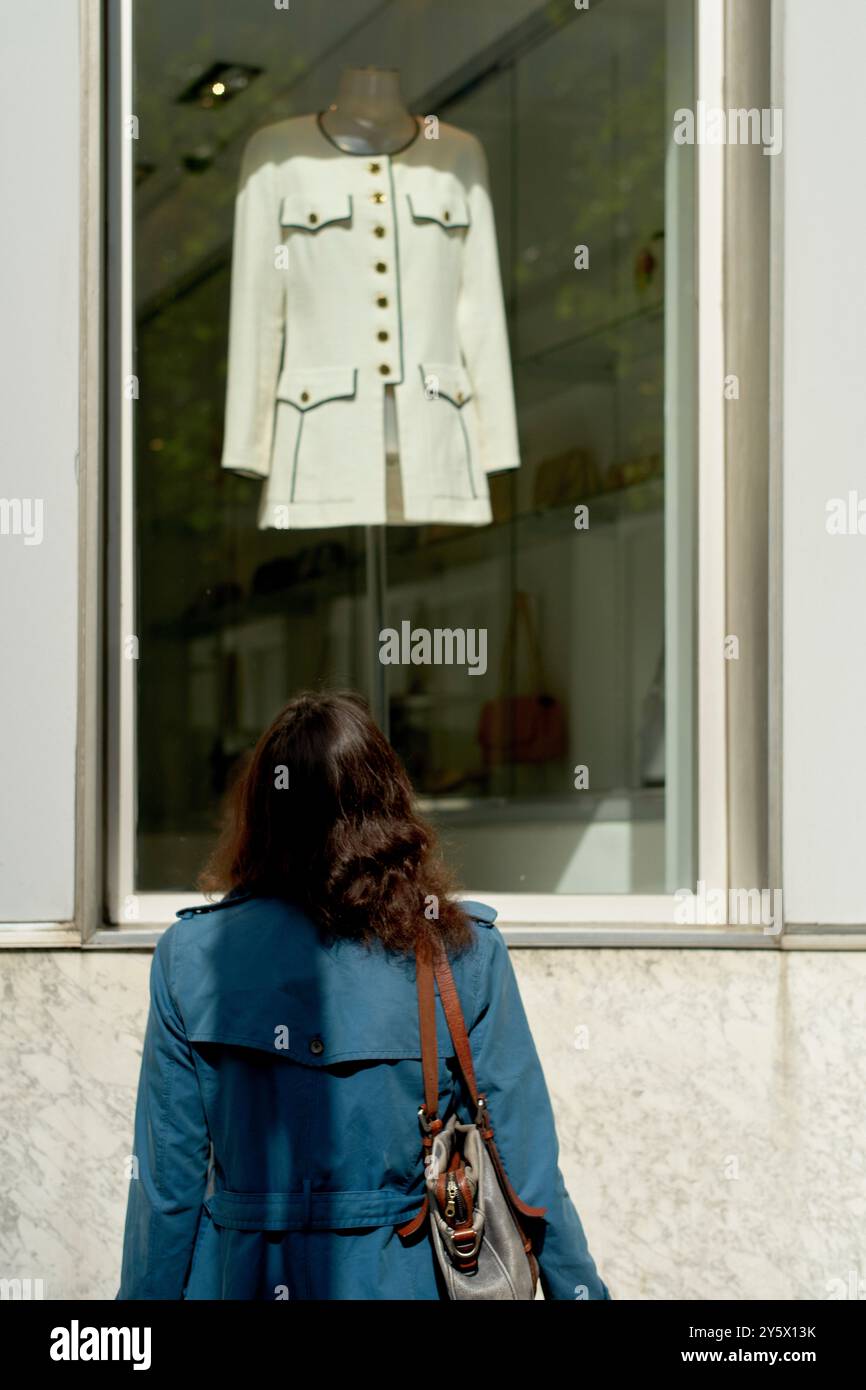 Una donna con un cappotto blu guarda una giacca bianca esposta in una vetrina. Foto Stock