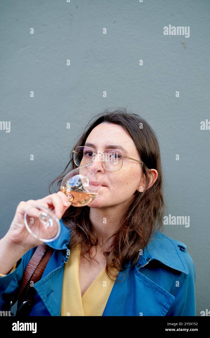 Donna con cappotto blu che sorseggia vino bianco su sfondo grigio. Foto Stock