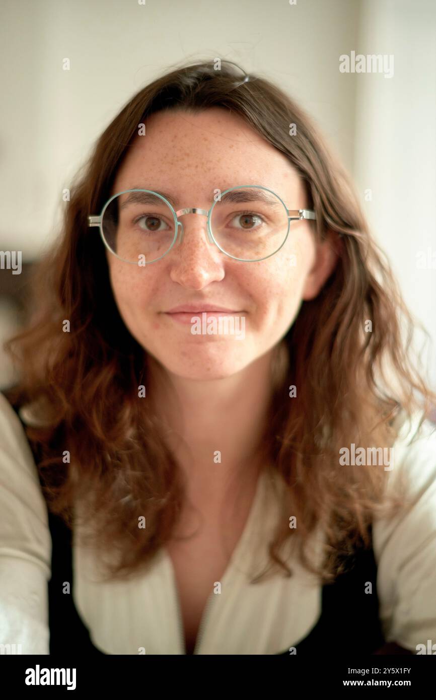 Giovane donna adulta sorridente con occhiali rotondi e capelli ondulati in una blusa bianca formale su uno sfondo morbido. Foto Stock