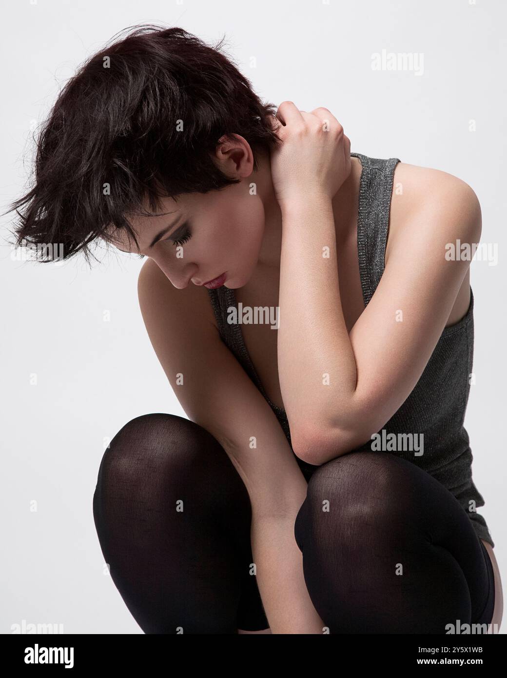 Giovane adulto con i capelli corti seduto in posizione accovacciata, tenendo le spalle, su uno sfondo bianco. Foto Stock