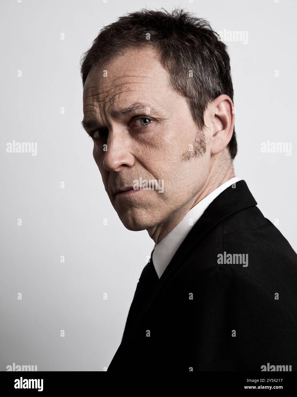 Primo piano di un serio uomo di mezza età con una leggera barba, vestito nero e camicia bianca, che guarda lateralmente su uno sfondo grigio. Foto Stock