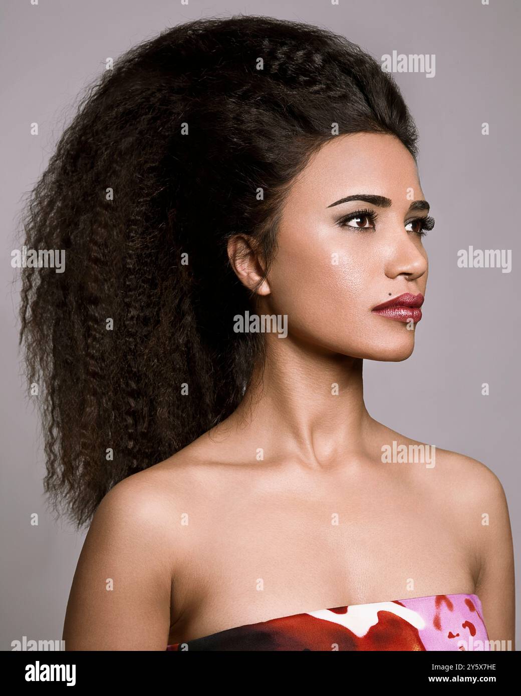 NA donna con voluminosi capelli ricci in posa con un abito colorato su sfondo grigio. Foto Stock NA donna con voluminosi capelli ricci in posa con un abito colorato su sfondo grigio. Foto Stock