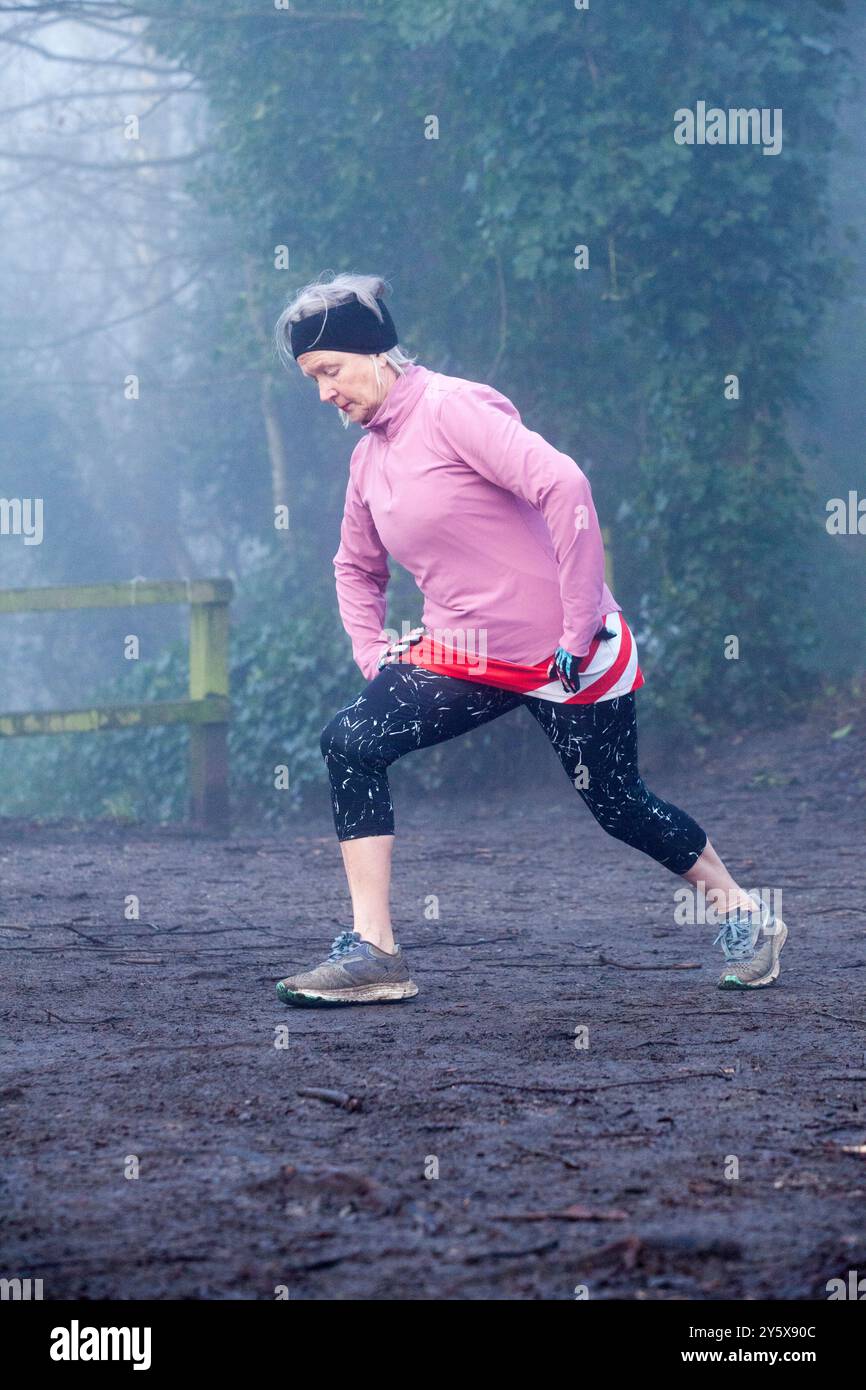 Donna anziana attiva che si allunga durante una corsa mattutina nebbiosa in un parco. Foto Stock