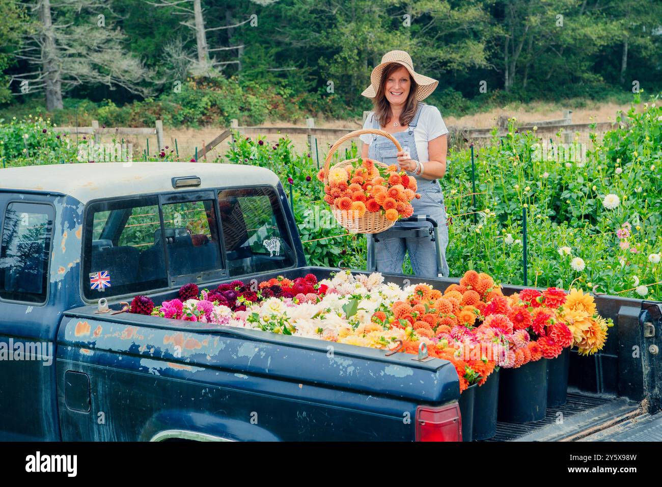 Donna sorridente in un cappello che tiene in mano un cesto di fiori accanto a un letto pieno di fiori colorati. Foto Stock