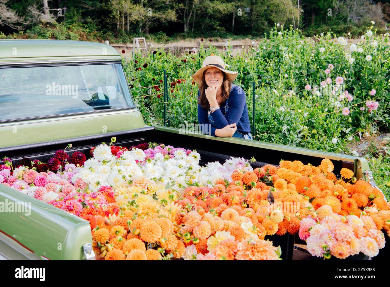 Donna sorridente con un cappello da sole si appoggia su un camion d'epoca pieno di fiori colorati in un giardino lussureggiante. Foto Stock