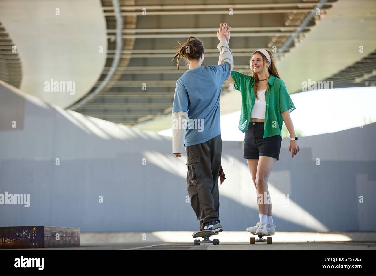 Due amici che condividono un alto cinque mentre fanno skateboard sotto un ponte, catturando un momento di gioia nella loro avventura urbana Foto Stock