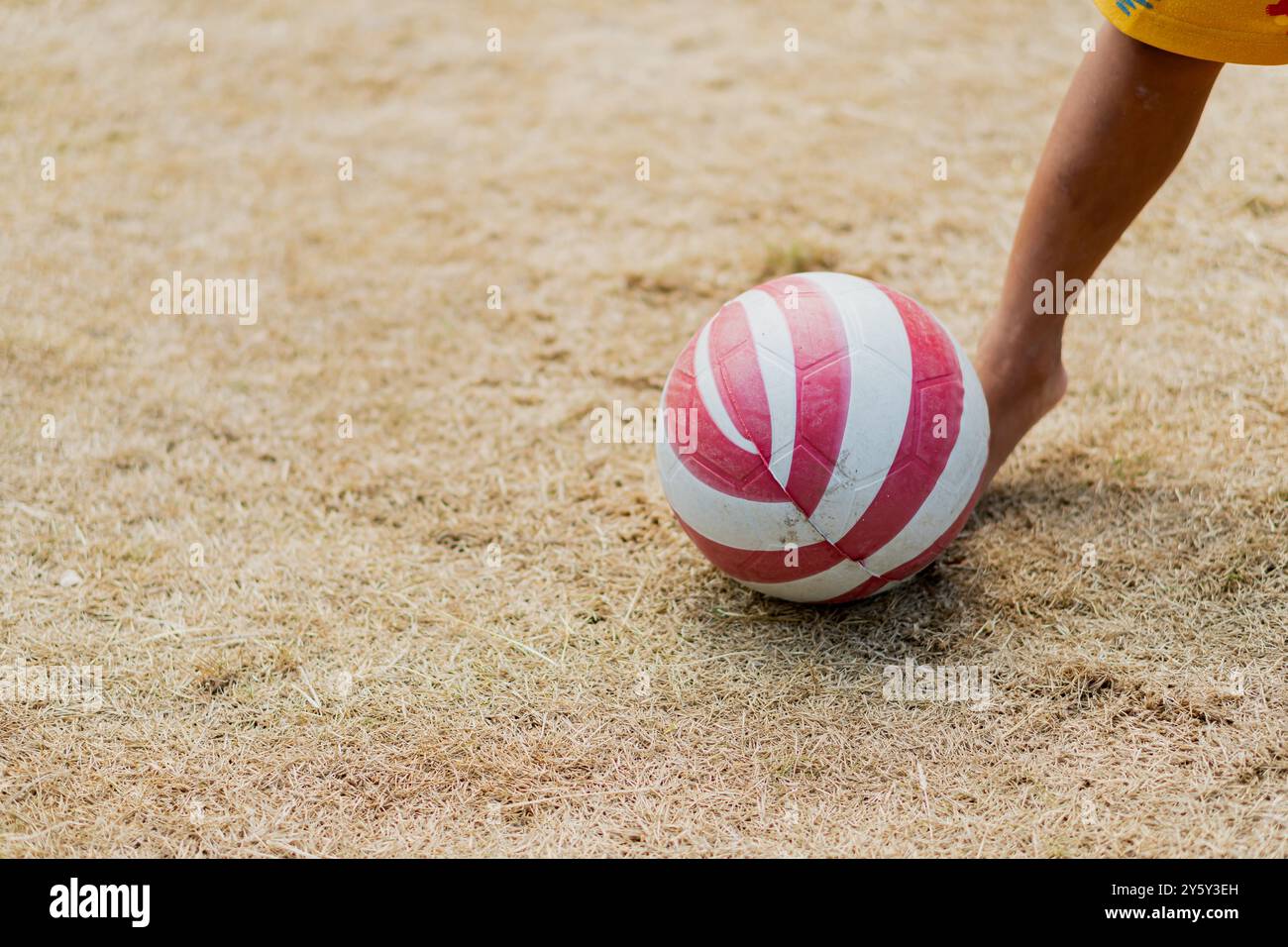 Primo piano di un bambino che calcia una palla a strisce rossa e bianca su un campo asciutto e erboso. La soleggiata scena all'aperto suggerisce giocosità e svago. Foto Stock