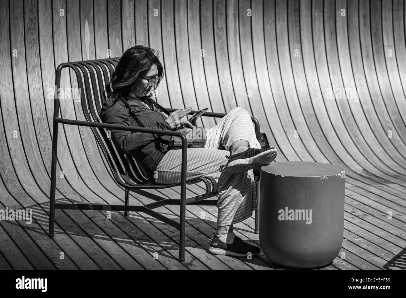 Donna asiatica in abiti casual e smartphone in mano seduta all'aperto nel parco urbano della città. Una donna siede sulla panchina usando il telefono cellulare. La gente si agita Foto Stock
