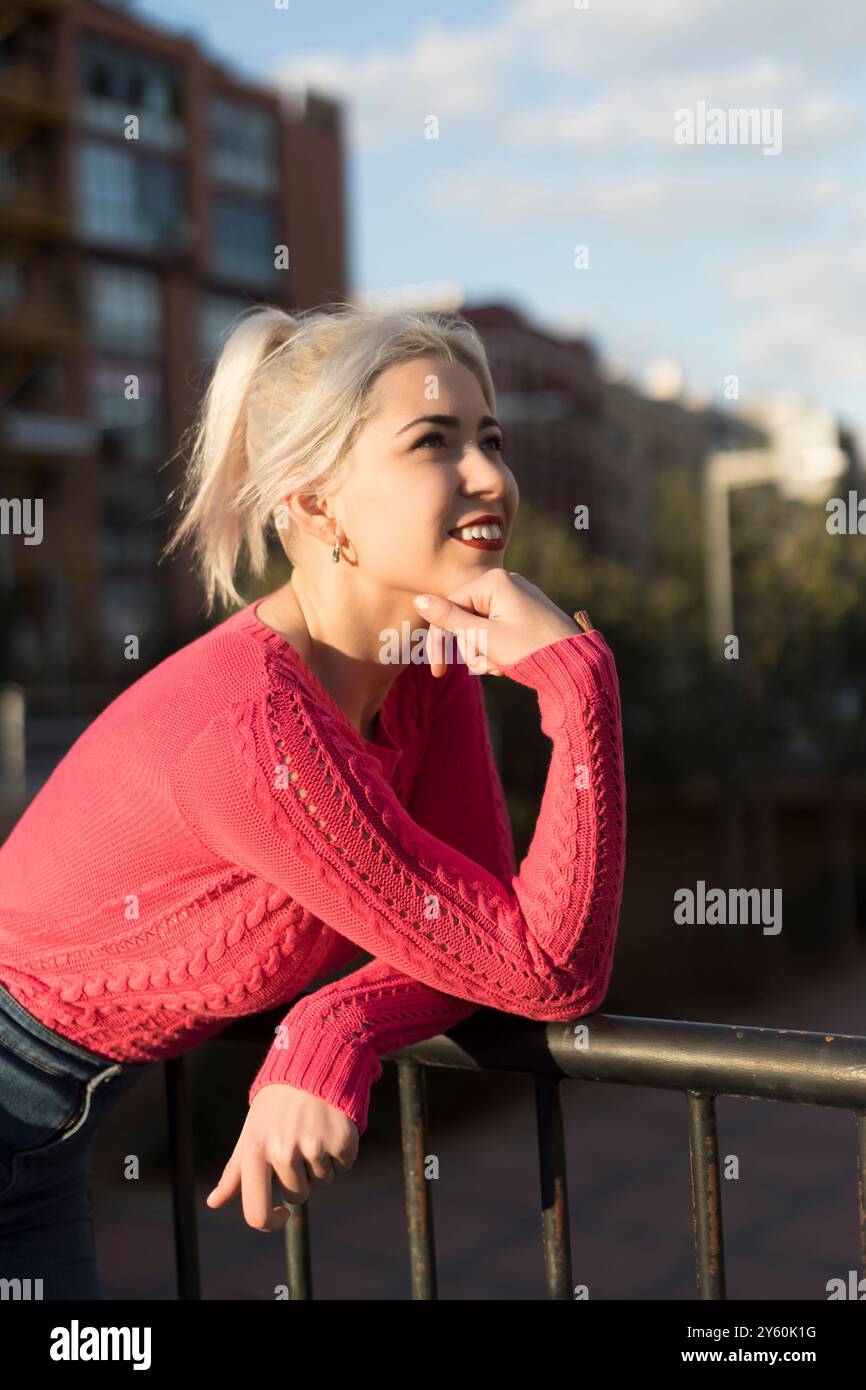 Una giovane donna con i capelli biondi platino sorride mentre si appoggia su una ringhiera, vestita con un maglione rosso, ammirando il tramonto in un ambiente urbano Foto Stock