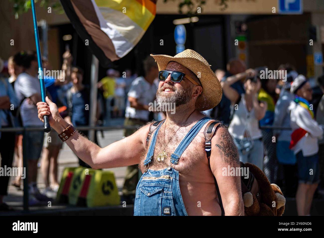Uomo barbuto con occhiali da sole, cappello da cowboy in paglia e tute in denim che sventolano bandiera con orso alla parata Helsinki Pride 2024 a Helsinki, Finlandia Foto Stock