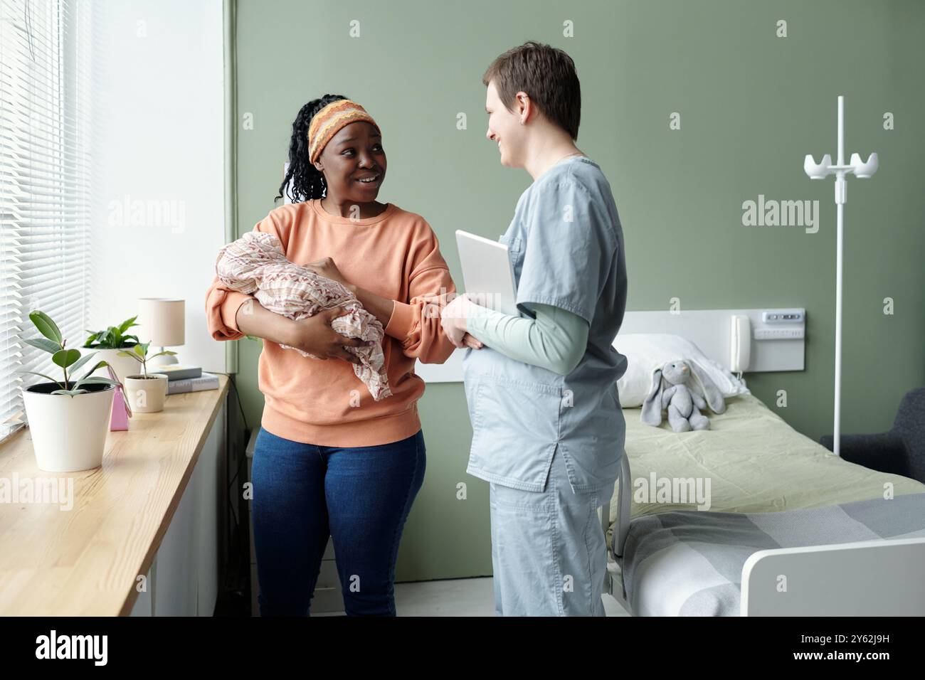 Madre sorridente che tiene in braccio il neonato mentre parla con un professionista sanitario in una stanza d'ospedale. La scena include piante in vaso e un letto d'ospedale Foto Stock
