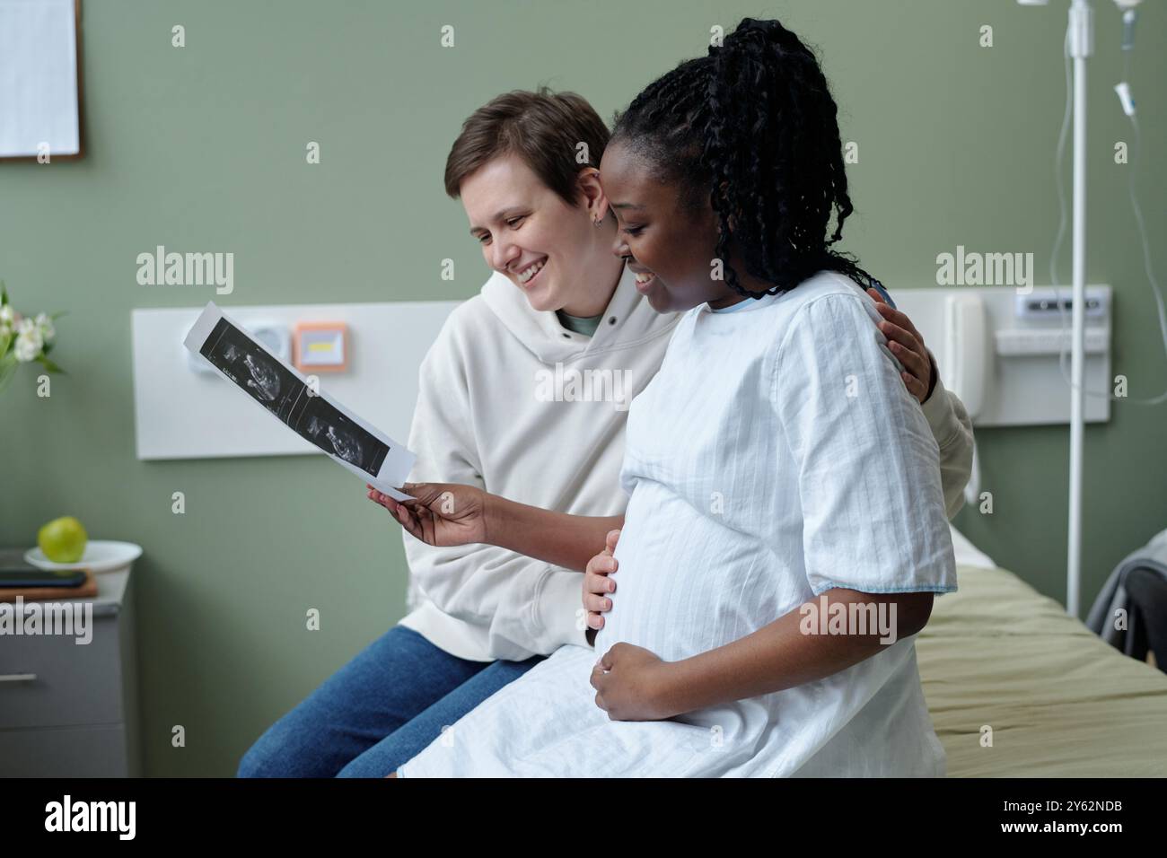 Donna incinta in abito ospedaliero che condivide l'immagine a ultrasuoni con un'altra persona sorridente in ambiente di stanza d'ospedale. Insieme, esprimono felicità e anticipazione Foto Stock