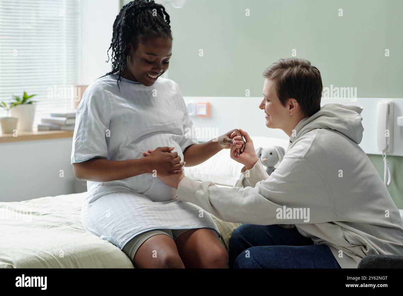 Donna incinta seduta sul letto d'ospedale con un'amica di supporto che tiene la mano, fornendo comfort e rassicurazione durante il momento potenzialmente stressante, al chiuso con attrezzature mediche visibili Foto Stock