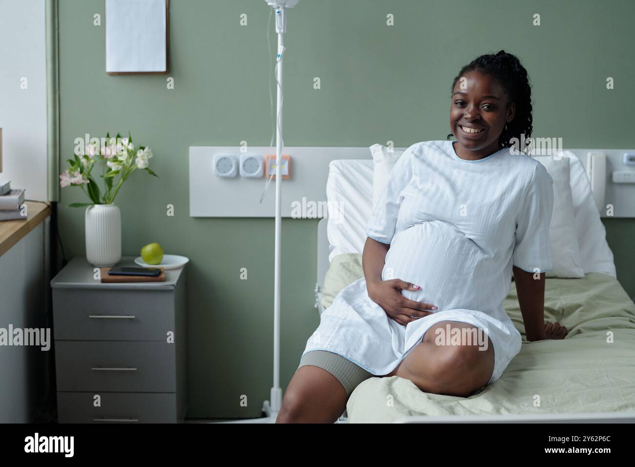 Donna afroamericana seduta nel letto d'ospedale mentre sorride e culla lo stomaco, con flebo IV attaccato al braccio e attrezzature mediche in background Foto Stock