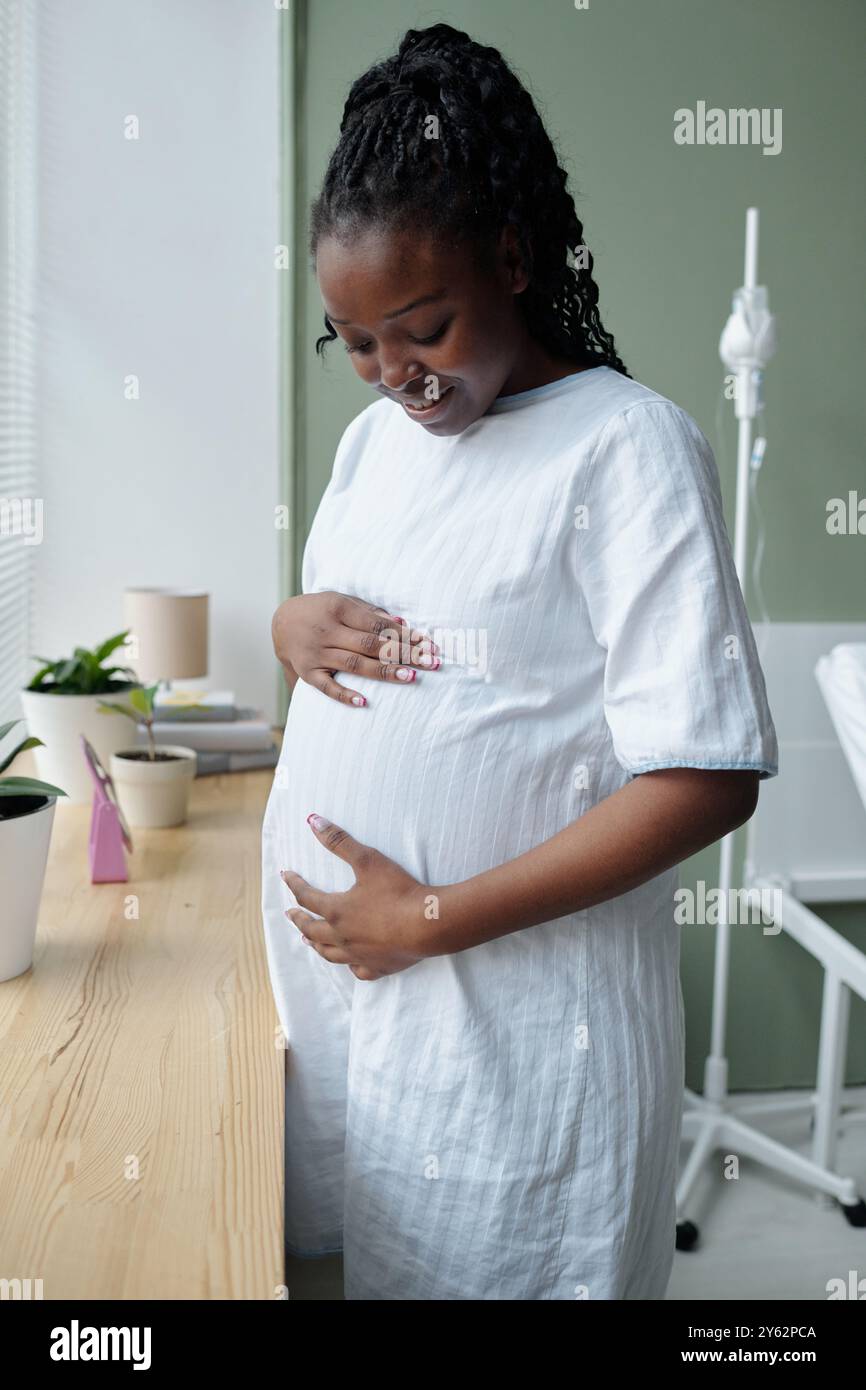 Donna afroamericana in piedi in abito bianco con la mano sulla pancia, guardando in basso, in un ambiente medico con piante in vaso e attrezzature mediche evidenti. Emorragia di luce materna Foto Stock