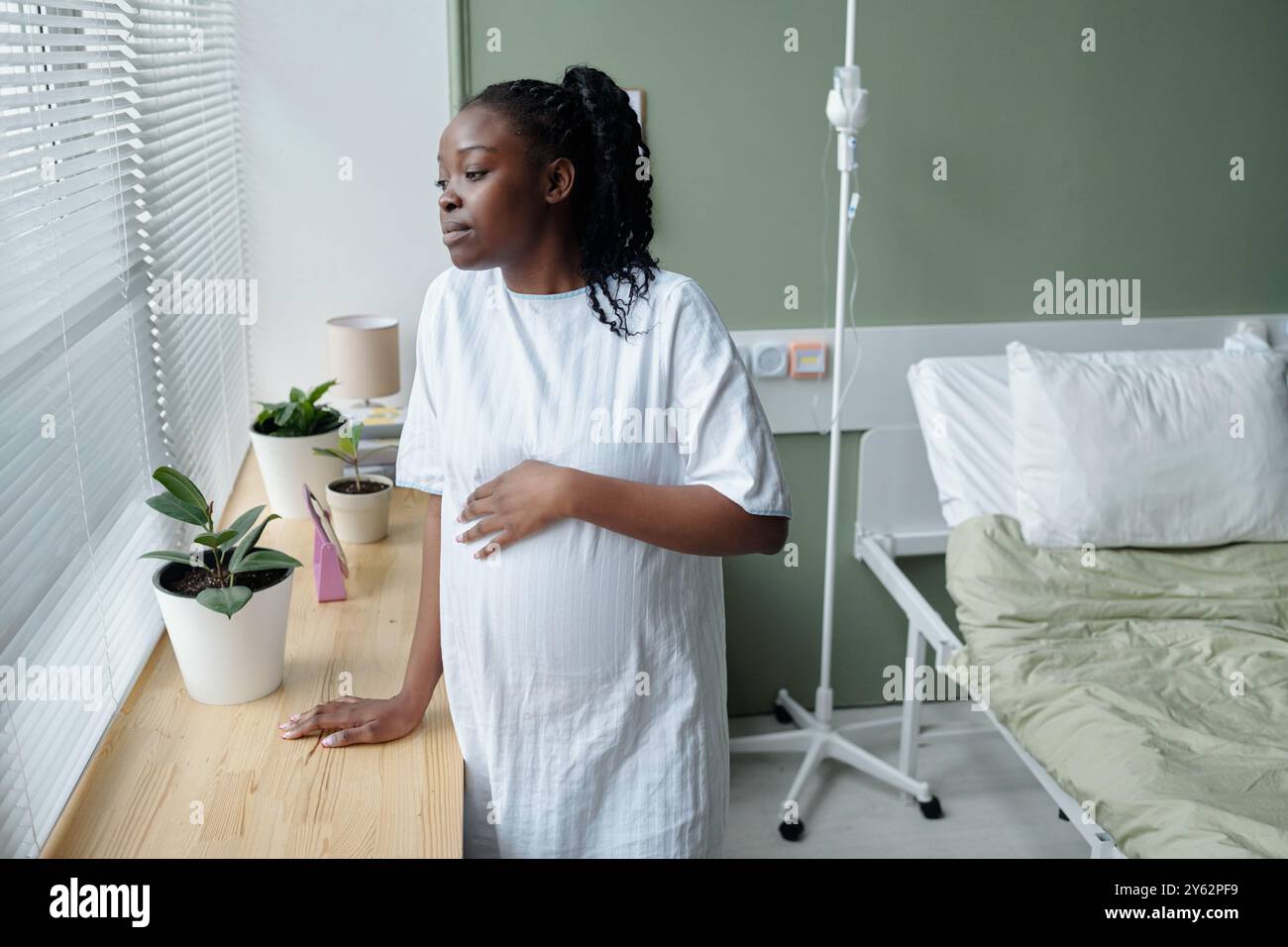 Donna incinta in piedi accanto a una finestra dell'ospedale tiene la pancia e guarda fuori. Letto d'ospedale e piante in vaso sullo sfondo creando un ambiente rilassante Foto Stock