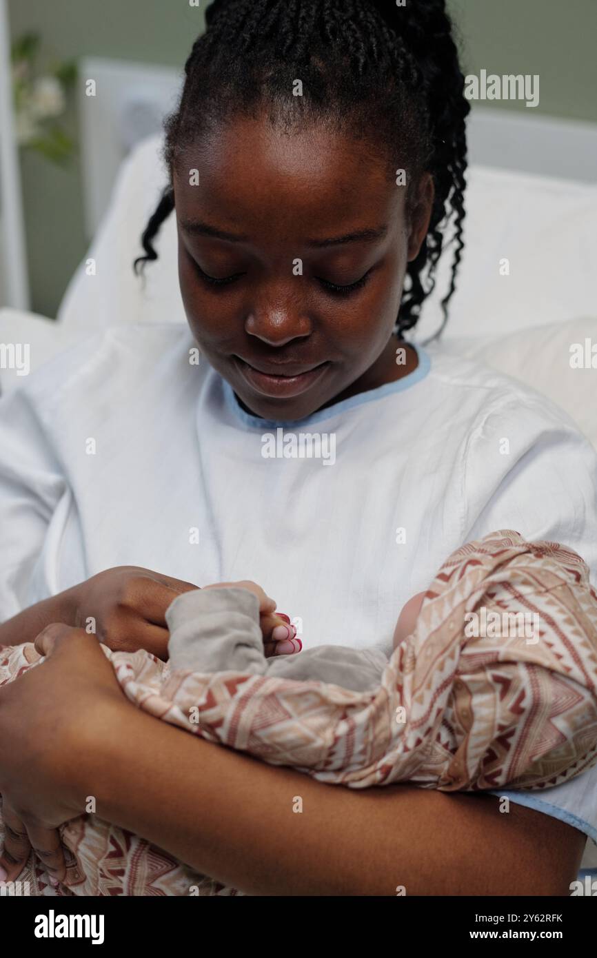 La giovane madre abbraccia il neonato addormentato nella stanza dell'ospedale, mostrando un'espressione calma e amorevole mentre tiene stretto. Un'illuminazione soffusa migliora la tranquillità e l'intimità Foto Stock
