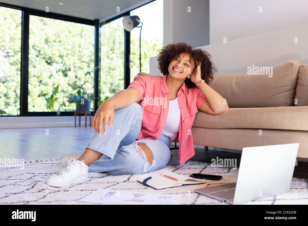 Donna sorridente seduta a terra con un notebook, un notebook e uno smartphone nelle vicinanze, a casa Foto Stock