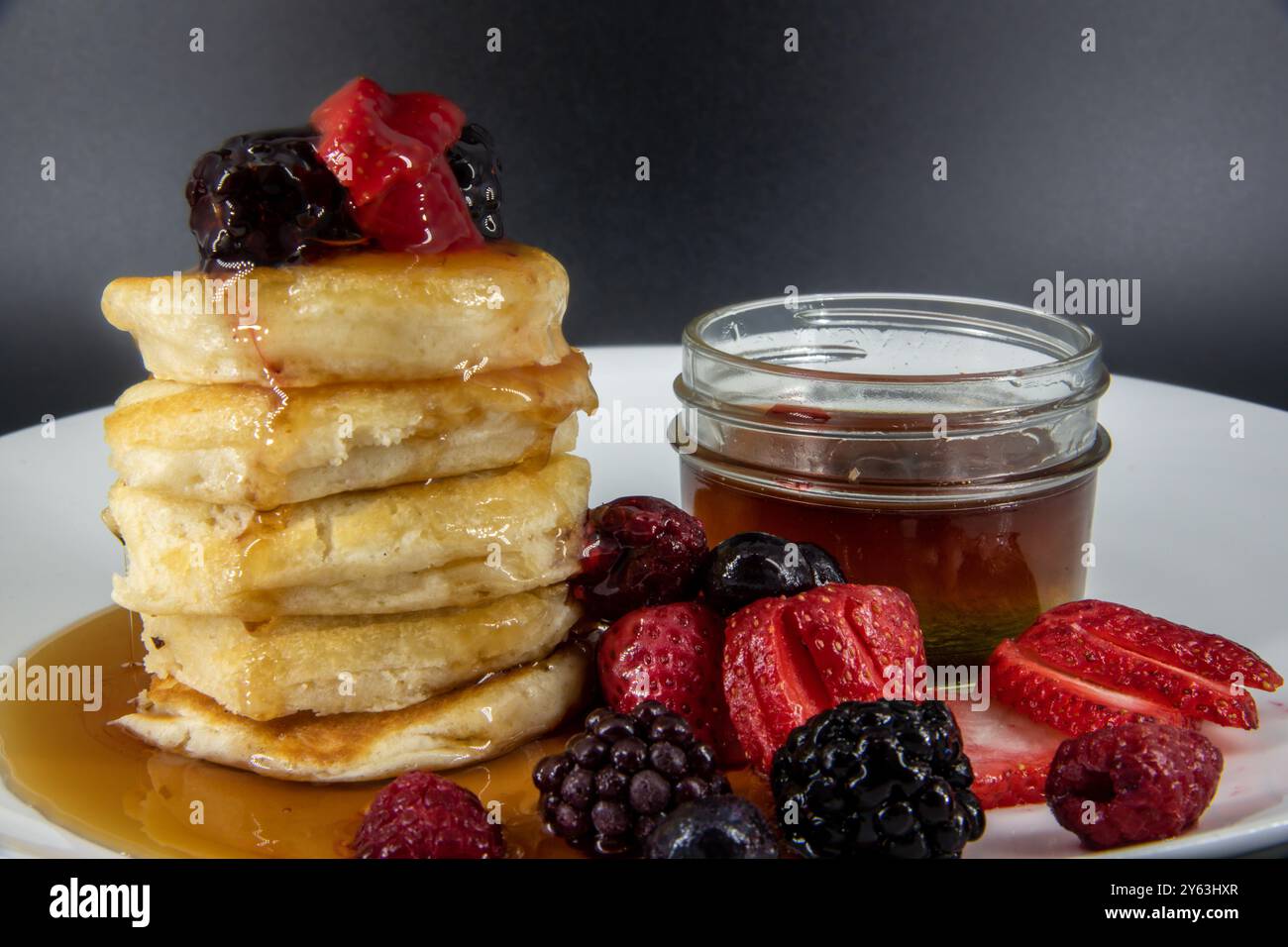 Gustosi pancake vegani e vegetariani al burro senza latticini con condimenti di bacche, sciroppo d'acero su un piatto bianco con sfondo nero. Foto Stock