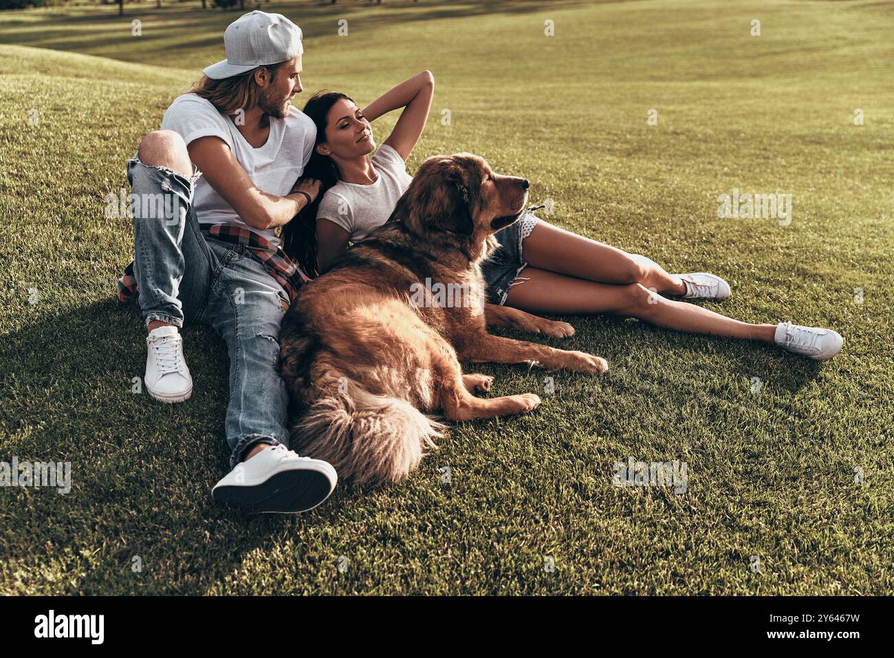 Vivere al momento. Giovane coppia moderna sdraiata sull'erba con il proprio cane mentre trascorre del tempo libero nel parco Foto Stock