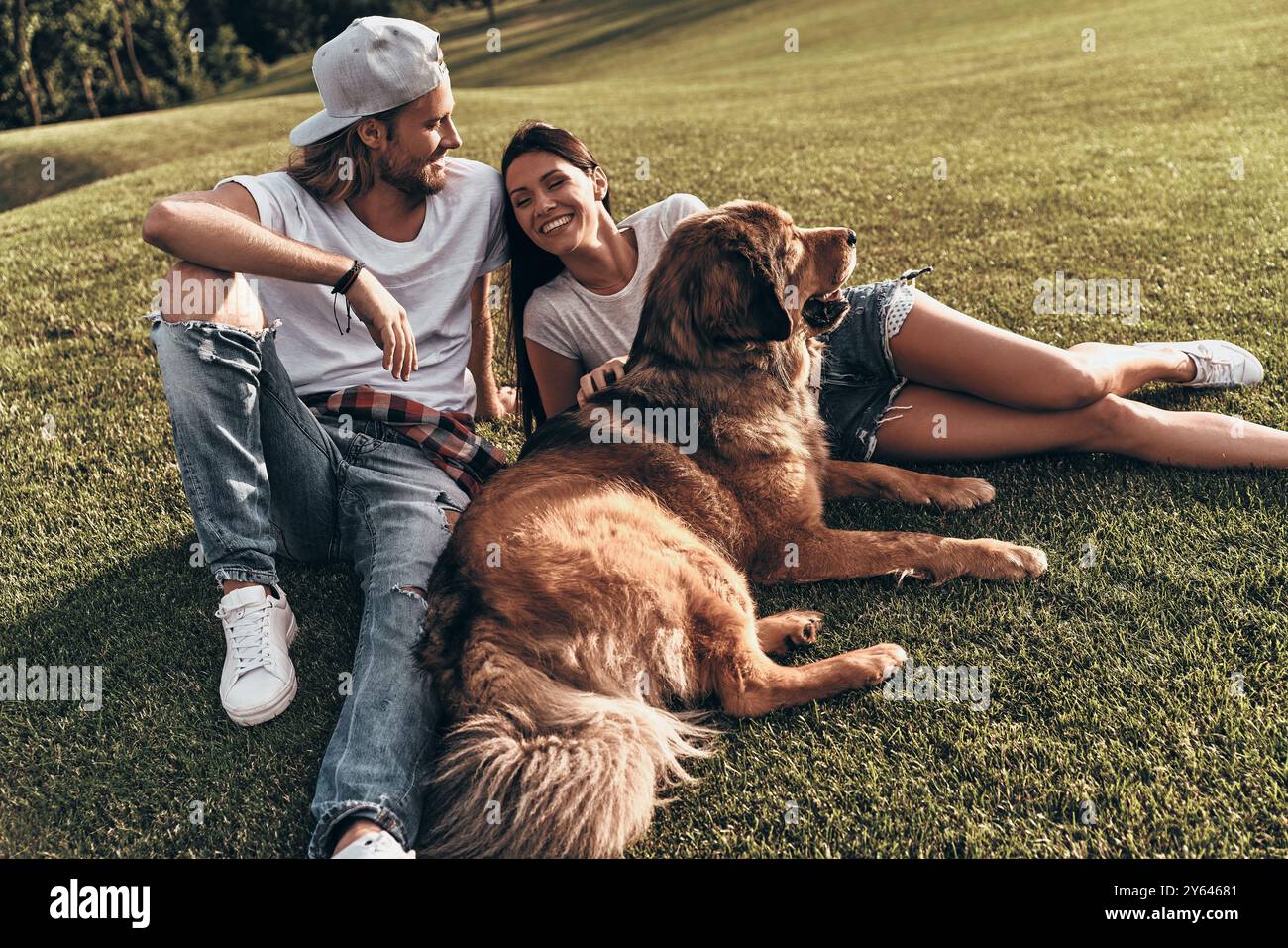 Buon tempo insieme. Giovane coppia moderna sdraiata sull'erba con il proprio cane mentre trascorre del tempo libero nel parco Foto Stock