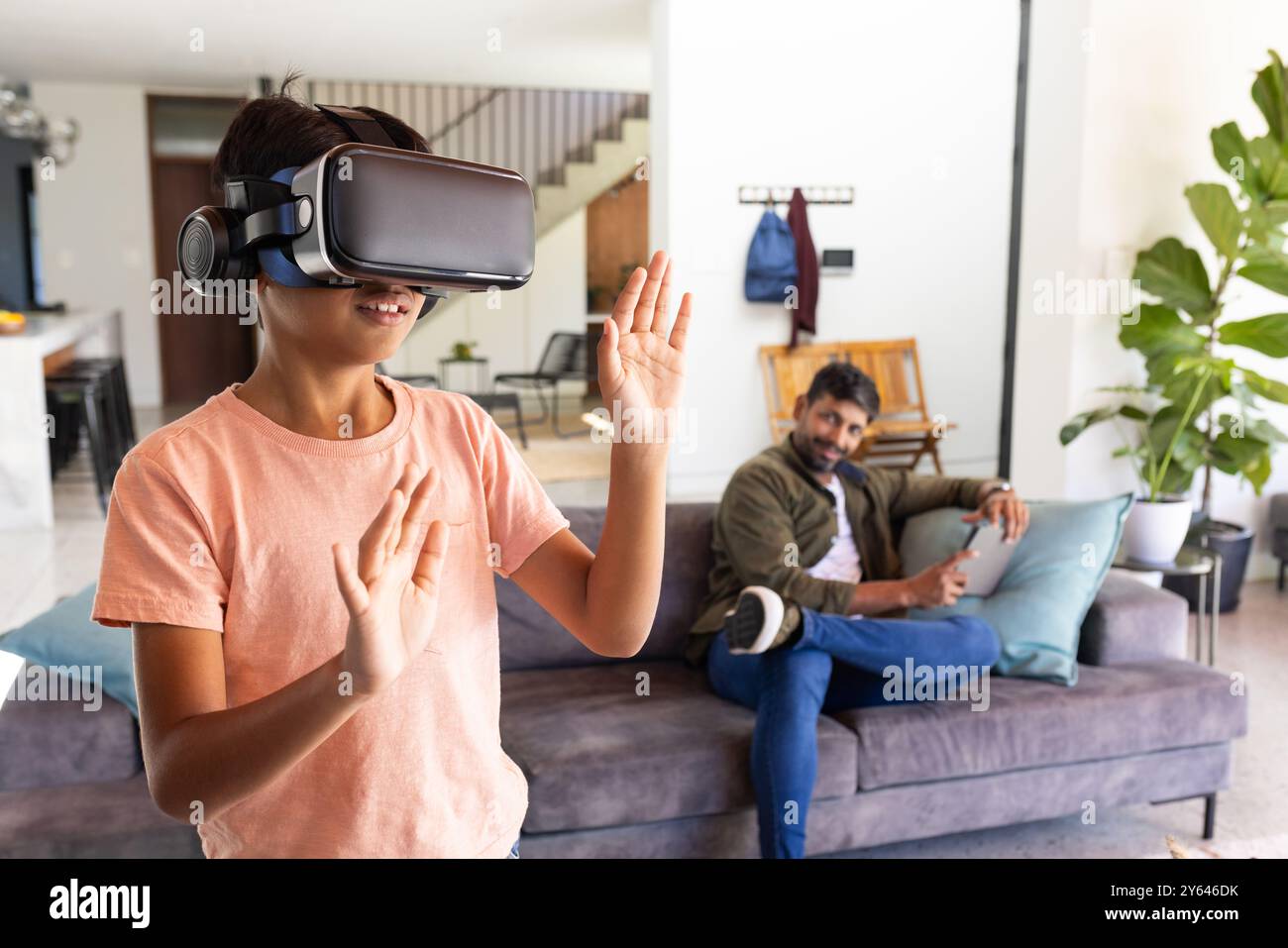 Utilizzando il visore VR, un ragazzo indiano che esplora il mondo virtuale mentre il padre indiano guarda sul divano Foto Stock