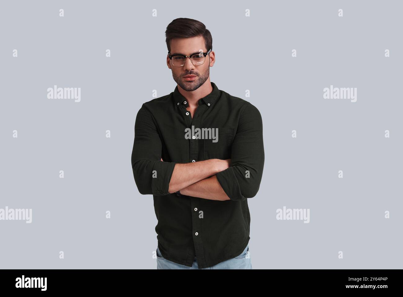 Aspetto serio. Giovane uomo con gli occhiali da vista che tiene le braccia incrociate e guarda la fotocamera mentre si trova su sfondo grigio Foto Stock