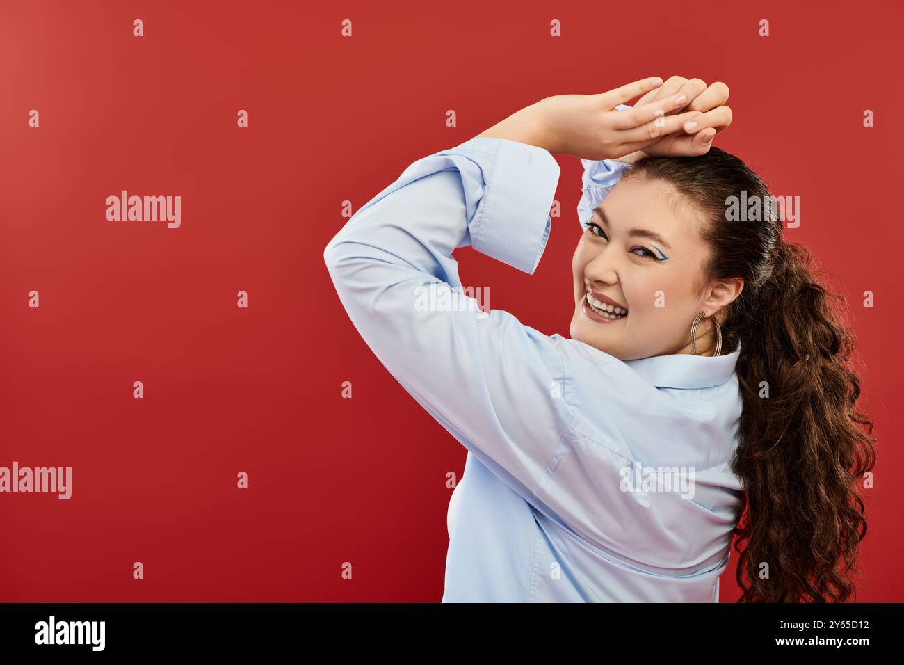 Una giovane donna di grandi dimensioni con un trucco luminoso posa allegramente con i capelli tirati indietro in un ambiente creativo da studio. Foto Stock