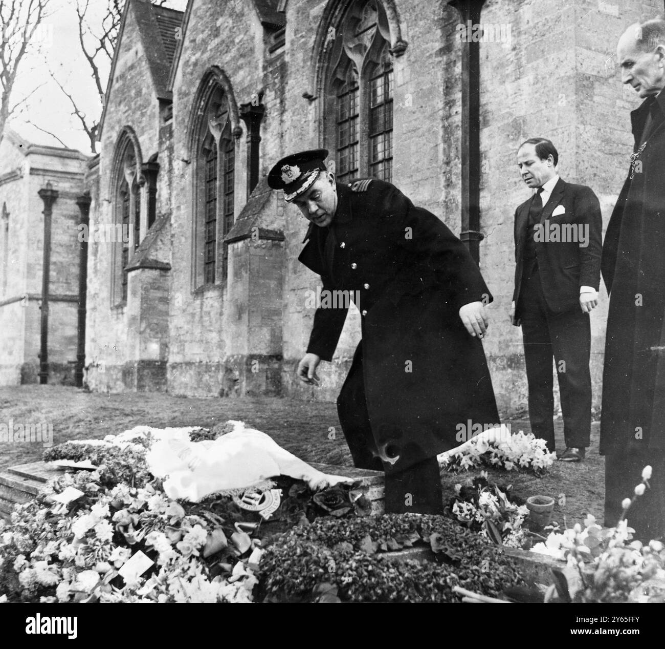 Ghirlanda greca per la tomba di Sir Winston Una ghirlanda contenente garofani e violette volate dalla Grecia fu posta nella tomba di Sir Winston Churchill per conto del governo greco dal capitano Spiridon Mourikis ( a sinistra ) , dalla Marina greca , dall' Esercito e dall' Aeronautica militare . A guardare vi sono il signor George Lyruoudias ( sfondo centrale ) funzionario dell' ambasciata a Londra e il Rettore di Woodstock e Bladon , il reverendo John e James che sabato ha condotto la cerimonia sulla tomba . 2 febbraio 1965 Foto Stock