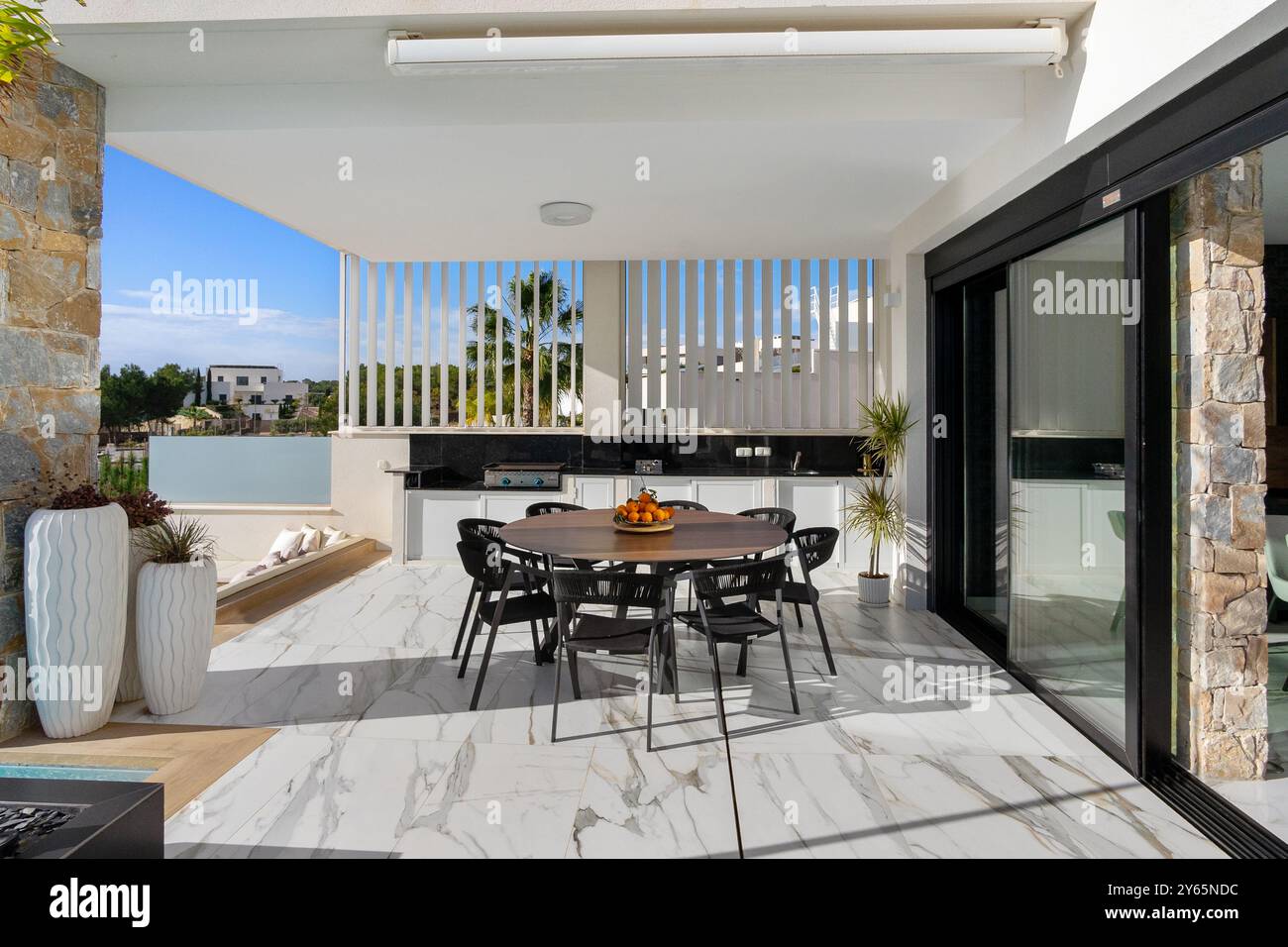 Questa villa moderna dispone di un'elegante cucina all'aperto e di una zona pranzo, completa di eleganti mobili neri e pavimento in marmo, con vista su un tranquillo ambiente Foto Stock