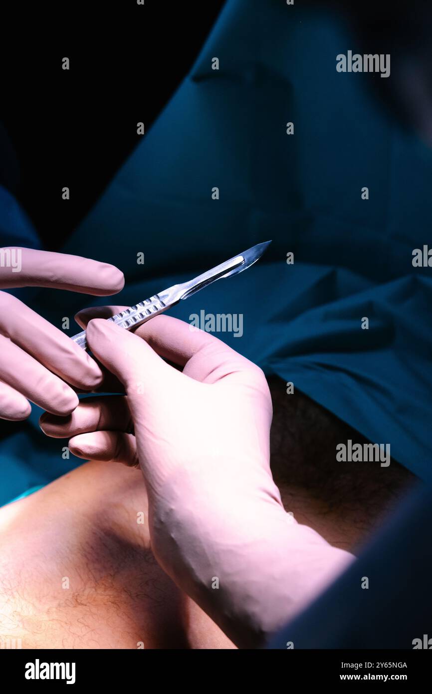 Vista dettagliata di un bisturi chirurgico tenuto da un medico durante un intervento chirurgico, con particolare attenzione allo strumento rispetto a un ambiente chirurgico sfocato Foto Stock