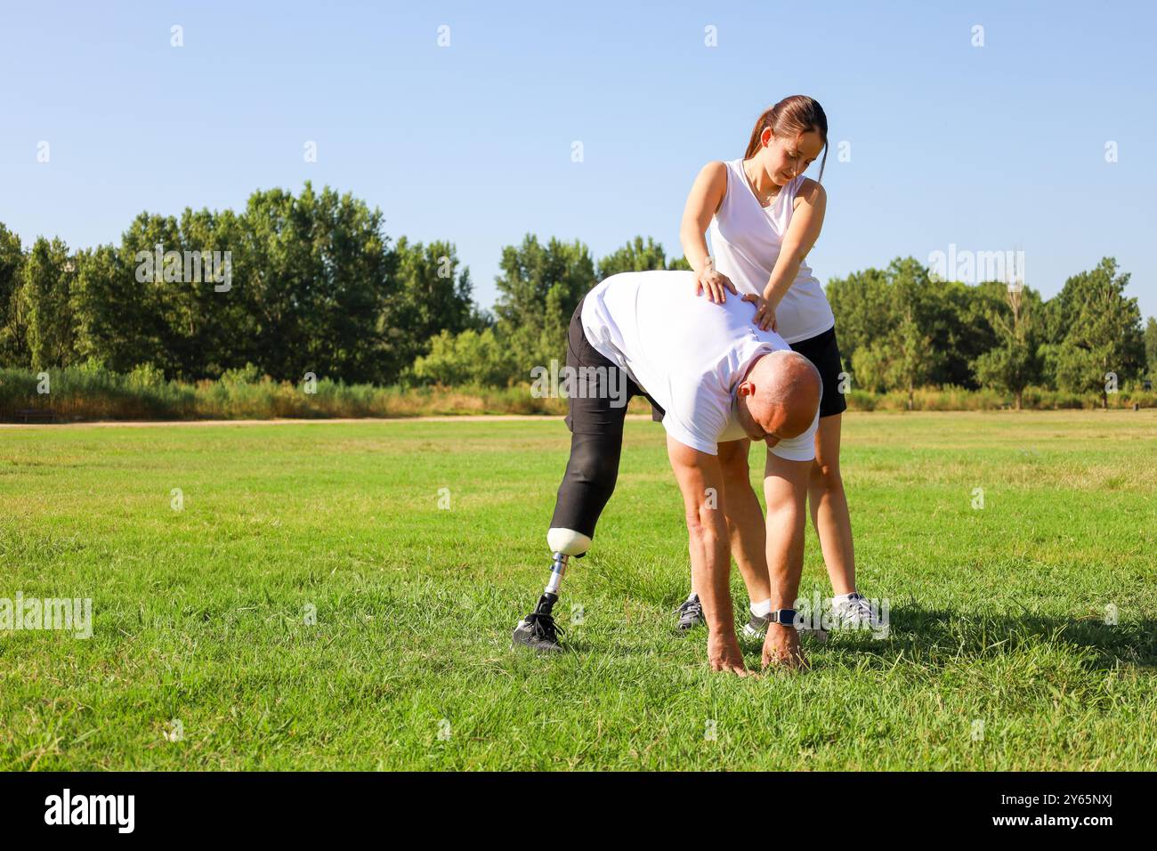 In un parco soleggiato, una figlia assiste suo padre, che ha una protesi della gamba, con esercizi di stretching, evidenziando un momento di sostegno e adattamento i Foto Stock