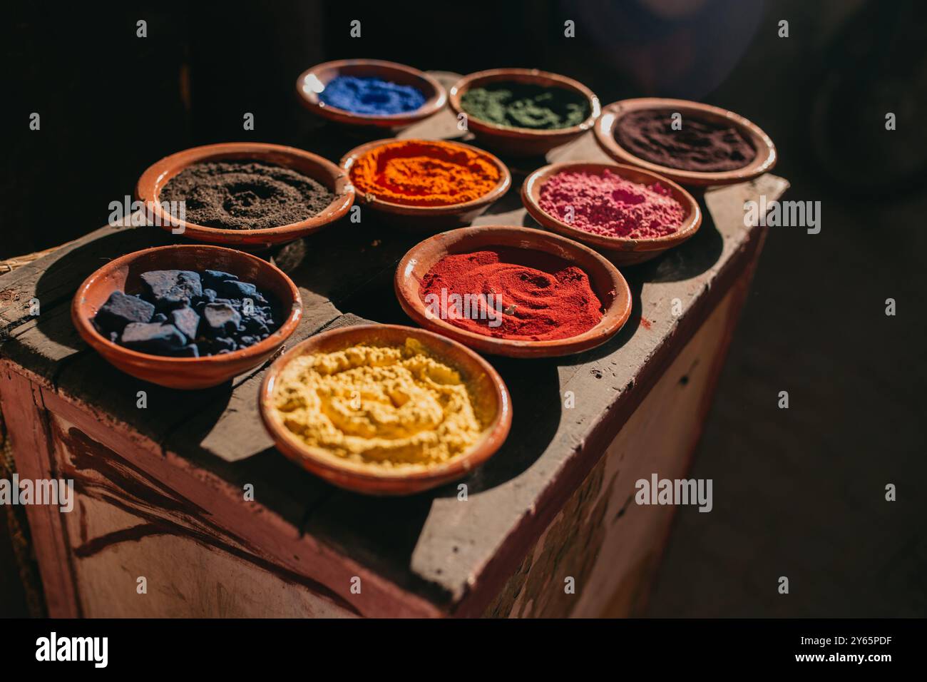 Vibranti spezie assortite in ciotole di terracotta poste su un tavolo di legno, illuminate dalla luce naturale del sole, raffiguranti un tradizionale mercato delle spezie a Marrakech Foto Stock