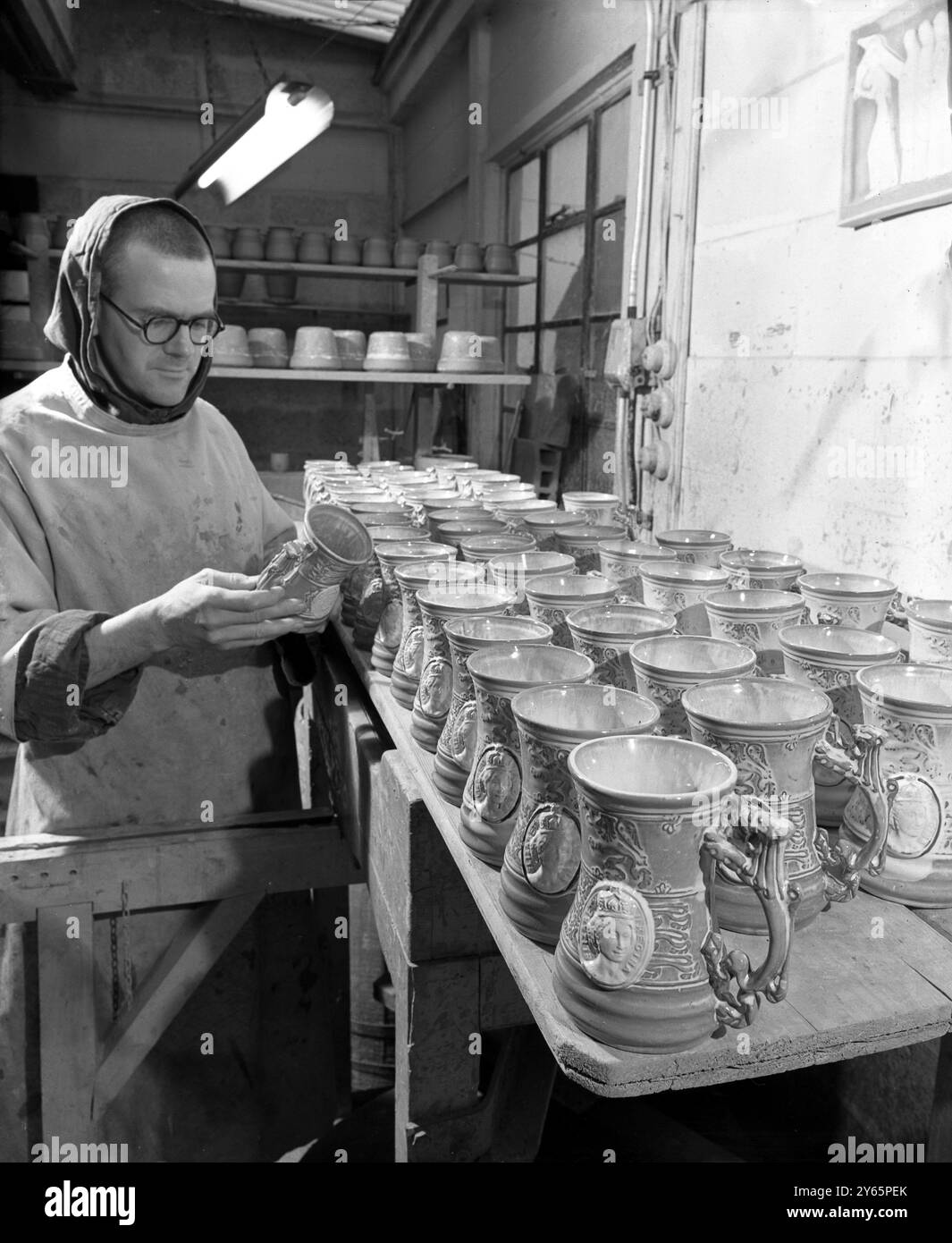 Monk fa i monaci dell'incoronazione. Fratello Thomas che ispezionava le tazze dell'incoronazione completate presso l'abbazia di Prinknash , Painswick, Gloucestershire , dove i monaci lavorano duramente nelle loro ceramiche per la produzione di questi e di altri souvenir dell'incoronazione . I monaci usano l'argilla scavata dal terreno dell'abbazia e lavorano su disegni originali. Ogni tazza - rifinita in verde , blu o crema - è decorata con il Royal Standard e una testa cammeo della Regina , con il monogramma ER , sul retro . Un leone rampante forma la maniglia . 13 dicembre 1952 Foto Stock