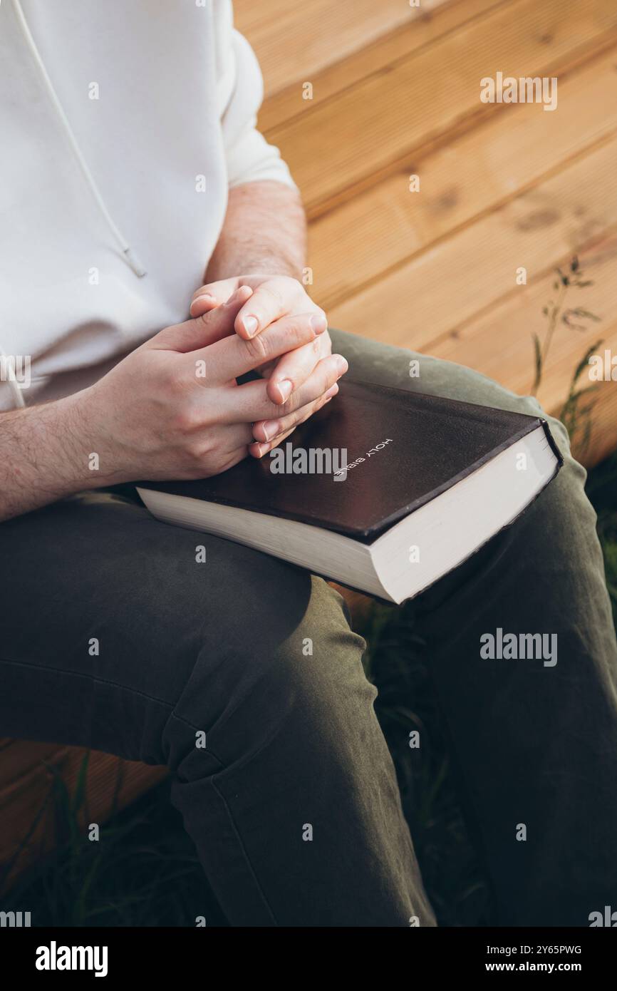 Vista ritagliata di un uomo irriconoscibile seduto all'aperto, che tiene e legge una Bibbia. Foto Stock