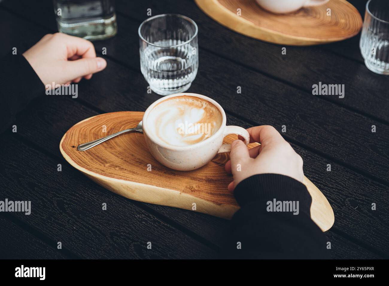 Vista dall'alto di un cappuccino appena preparato con un'elegante schiuma, servito su un piatto di legno a forma di cuore. La mano di una persona, tagliata e irriconoscibile, Foto Stock