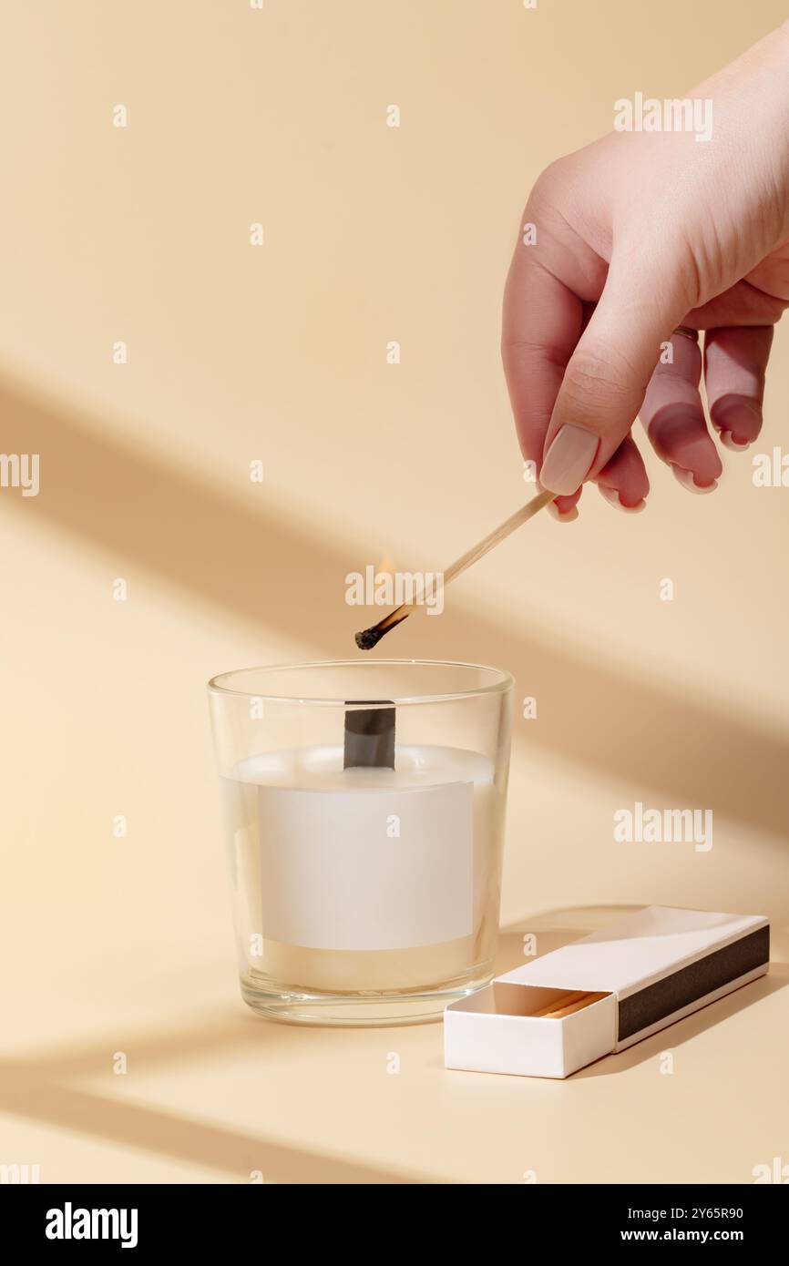 La mano di una persona viene catturata mentre accende una partita, pronta ad accendere una candela bianca profumata posizionata all'interno di un contenitore di vetro trasparente. La scena è ambientata a. Foto Stock