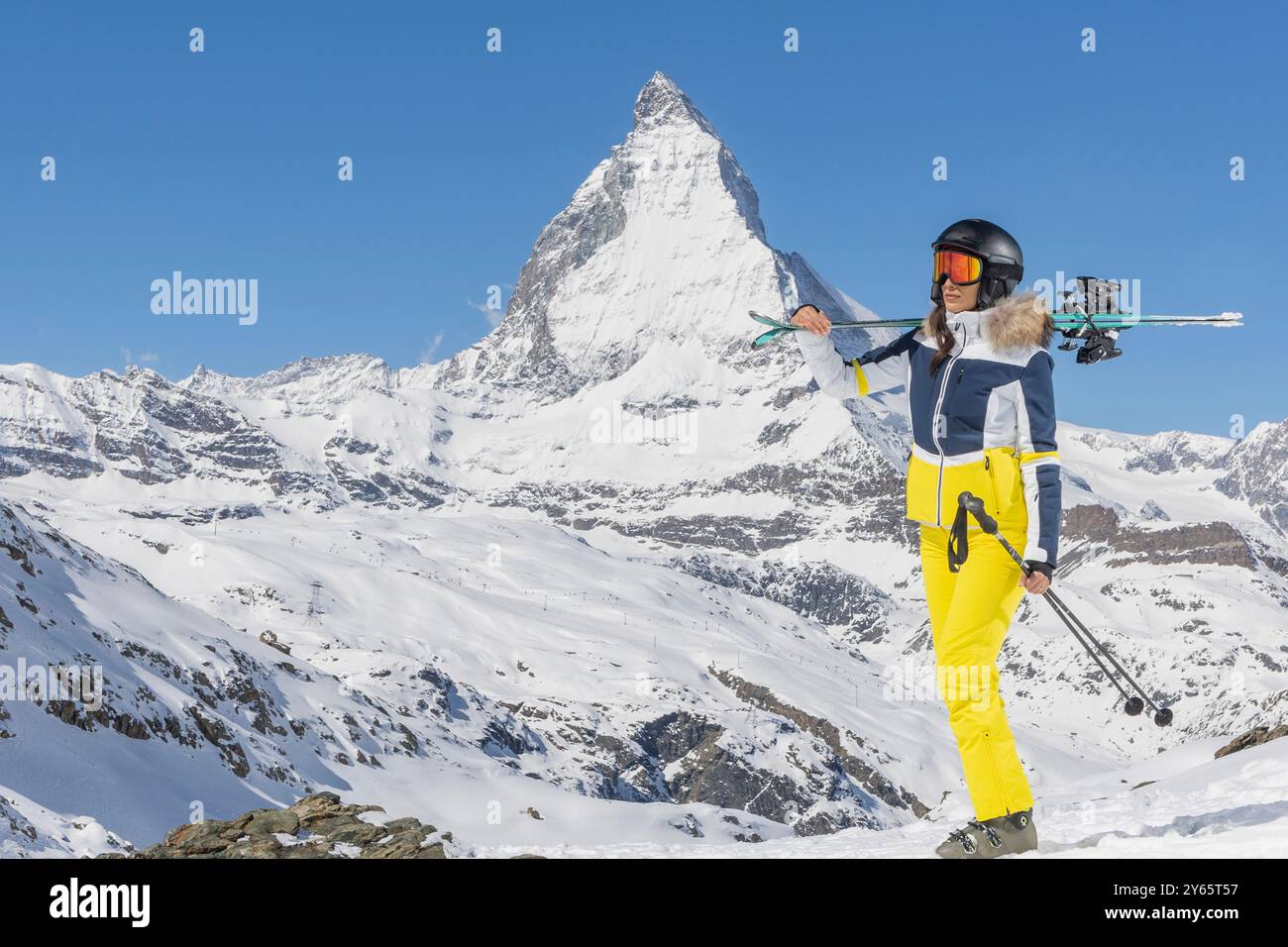 Di fronte al maestoso Cervino si trova una sciatrice con una moderna tuta gialla e blu, dotata di sci e pali, che incarnano moda e fu Foto Stock